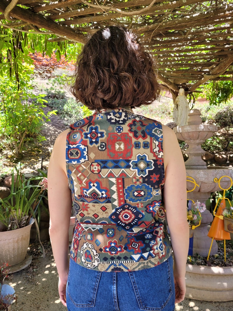 May include: A sleeveless vest with a geometric pattern in shades of brown, blue, red, and white. The vest is made of a woven fabric and has a relaxed fit.