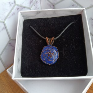 May include: A deep blue lapis lazuli stone pendant wrapped in thin copper wire, featuring a geometric wire pattern on the front and a coiled wire bail. The pendant is suspended from a simple black cord and rests inside a white gift box.