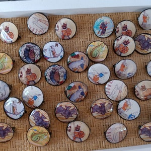 May include: A collection of round, antique-style brooches with various medieval illustrations. The brooches feature images of animals, figures, and text, set against a cream-colored background. Each brooch has a black metal frame and a pin on the back.