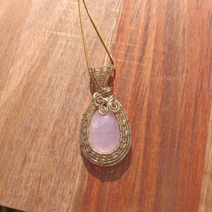 May include: A teardrop-shaped rose quartz pendant wrapped in gold-colored wire, featuring a woven lattice pattern around the stone and a decorative spiral accent at the top. The pendant hangs from a thin gold-colored chain necklace.