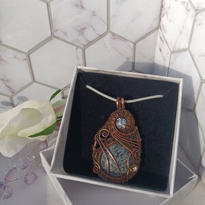 May include: A wire-wrapped pendant featuring a round, grey and black snowflake obsidian stone encased in oxidized copper wire, accented with two small clear faceted crystals, suspended on a light-colored cord inside a black-lined gift box.