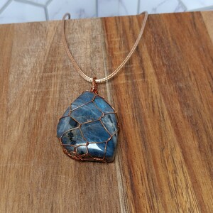 May include: A pendant featuring a polished, irregular blue gemstone encased in a decorative copper wire cage. The stone displays dark inclusions and iridescent blue tones. It is suspended from a thin, light-colored cord necklace.