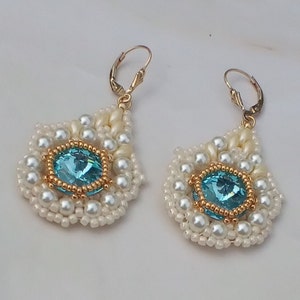 May include: A pair of drop earrings featuring a central round light blue crystal surrounded by a border of small gold beads. The crystals are framed by layers of white seed beads and white pearls, finished with gold-toned lever back ear wires.