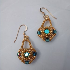 May include: A pair of dangle earrings featuring a central round teal crystal surrounded by metallic gold seed beads. Each earring has a geometric beaded frame accented with three dark teal faceted beads and hangs from a gold-toned wire hook.