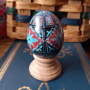 May include: A blue and red decorated Easter egg with a geometric pattern sits on a wooden stand. The egg is on a blue book with a gold design.