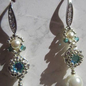 May include: A pair of drop earrings featuring silver-toned metal hooks embedded with clear crystals. Each earring consists of a central white pearl, small silver seed beads, and teal-colored crystal accents arranged in a circular pattern.