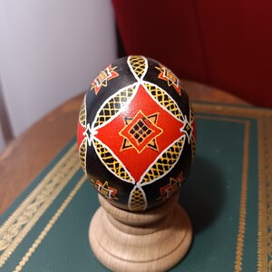 May include: A black, red, and yellow decorated Easter egg on a wooden stand. The egg has a geometric pattern with a diamond shape in the center.
