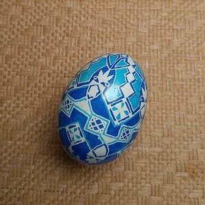 May include: A blue and white decorated Easter egg with a geometric pattern. The egg is painted with white lines and shapes on a blue background.
