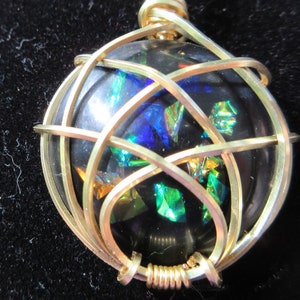 May include: A round black pendant featuring embedded iridescent shards in shades of blue, green, and orange. The circular stone is encased in a decorative gold-toned wire cage that wraps around the surface, secured with a coiled wire bail at the top.