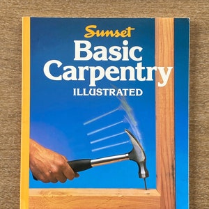 May include: A book titled "Sunset Basic Carpentry Illustrated" with a yellow spine and a blue cover featuring a hand holding a hammer hitting a nail into a piece of wood.