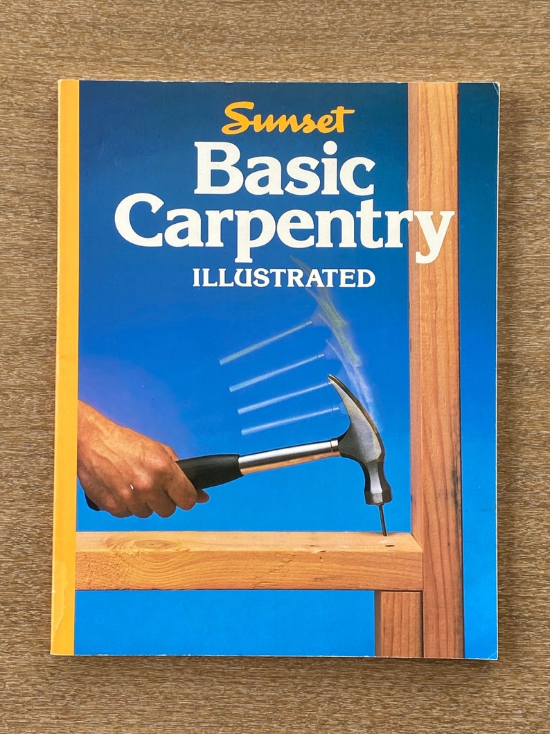 May include: A book titled "Sunset Basic Carpentry Illustrated" with a yellow spine and a blue cover featuring a hand holding a hammer hitting a nail into a piece of wood.
