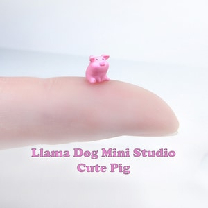 以下が含まれることがあります： 指の上に置かれた小さなピンクの豚の置物。置物は高さ約2.5cmで、可愛らしい漫画のような表情をしています。置物の下に「Llama Dog Mini Studio Cute Pig」という文字が見えます。