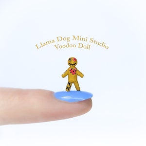 May include: A miniature yellow voodoo doll with black cross-stitch details and red heart and eye accents. The doll is standing on a blue surface. The text "Llama Dog Mini Studio Voodoo Doll" is in the background.