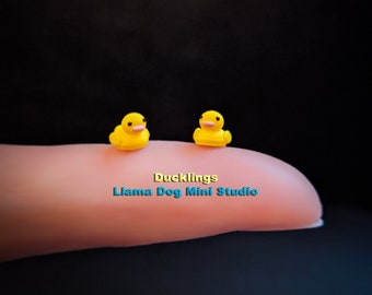 May include: Two small yellow rubber ducklings, one facing left and one facing right, on a finger. The text "Ducklings Llama Dog Mini Studio" is below the ducklings.