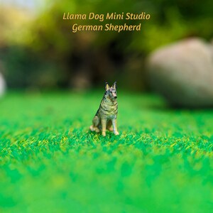Może przedstawiać: Miniaturowa figurka psa owczarka niemieckiego siedząca na zielonej trawie. Figurka jest wykonana z jasnobrązowego i szarego materiału, z czarnymi oczami i czarnym nosem.