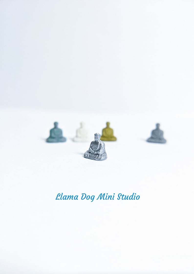 May include: A silver miniature Buddha statue sitting in a meditative pose. The statue is surrounded by four other miniature Buddha statues in different colours, including grey, white, and gold.