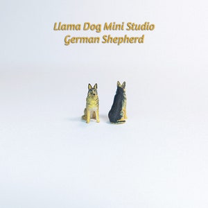 Może przedstawiać: Dwie miniaturowe figurki niemieckich owczark&oacute;w, jedna czarna i brązowa, druga brązowa i beżowa, siedzące na białym tle. Tekst "Llama Dog Mini Studio German Shepherd" znajduje się nad figurkami.