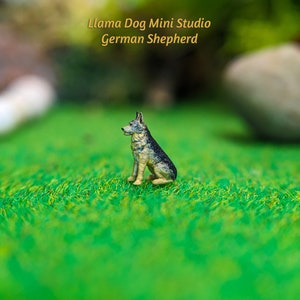 Może przedstawiać: Miniaturowa figurka psa rasy owczarek niemiecki, siedząca na zielonej trawie. Figurka jest wykonana z malowanego metalu i ma realistyczny wygląd. Tekst "Llama Dog Mini Studio German Shepherd" jest widoczny nad figurką.