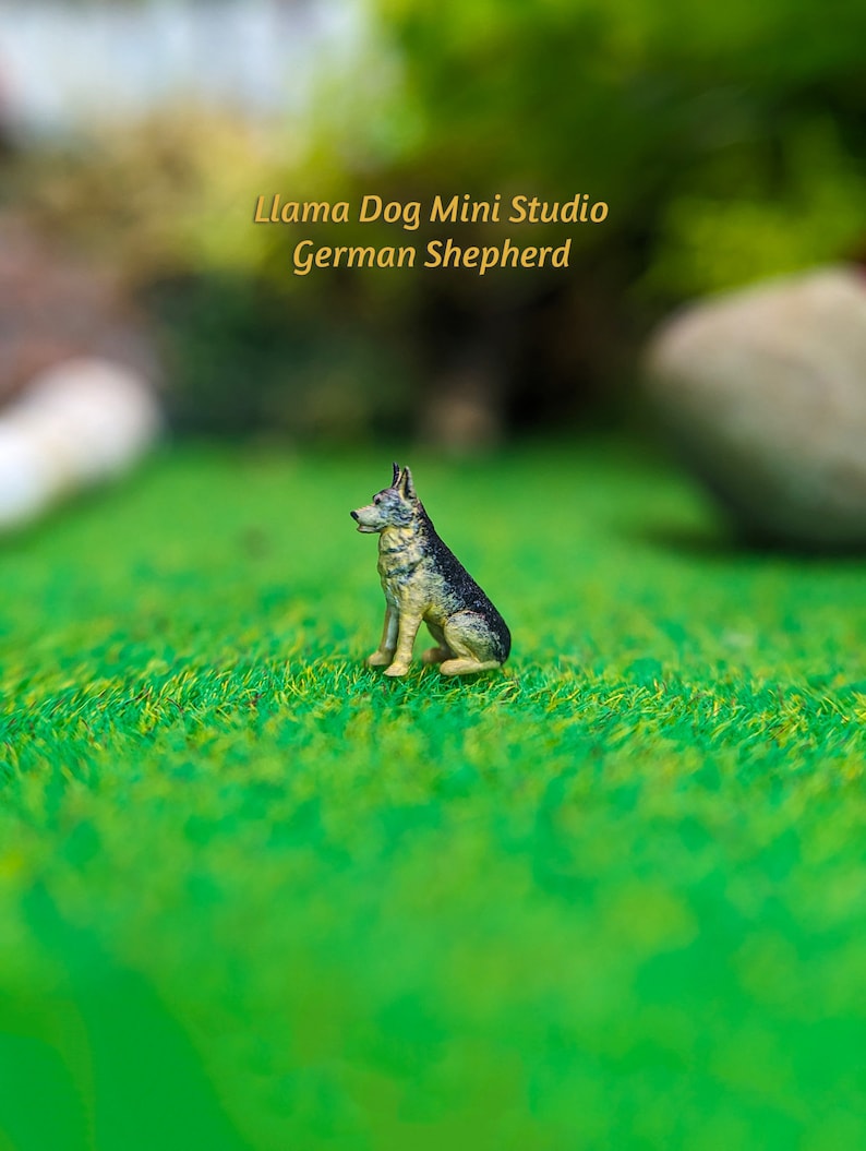 Może przedstawiać: Miniaturowa figurka psa rasy owczarek niemiecki, siedząca na zielonej trawie. Figurka jest wykonana z malowanego metalu i ma realistyczny wygląd. Tekst "Llama Dog Mini Studio German Shepherd" jest widoczny nad figurką.