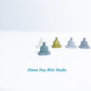 May include: Five miniature figurines of Buddha in various poses, sitting on a white surface. The Buddha in the centre is green, while the others are white, gold, and grey.