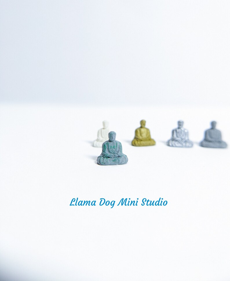 May include: Five miniature figurines of Buddha in various poses, sitting on a white surface. The Buddha in the centre is green, while the others are white, gold, and grey.
