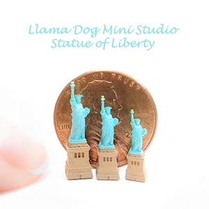 May include: Three miniature Statue of Liberty replicas, each a different size, standing on a base. The statues are turquoise blue and the bases are tan. A US penny is in the background for scale.