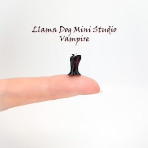 May include: A miniature vampire figurine, black with a red-lined cape, perched on a fingertip. The figurine is part of the Llama Dog Mini Studio collection. The word "Vampire" is written below the studio name.
