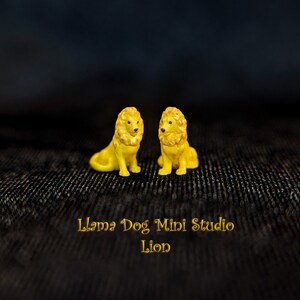 K&ouml;nnte beinhalten: Zwei kleine gelbe L&ouml;wenfiguren, die sich gegen&uuml;berstehen. Die Figuren sitzen auf einer schwarzen, strukturierten Oberfl&auml;che. Der Text "Llama Dog Mini Studio Lion" befindet sich unter den Figuren.