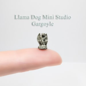 May include: A small, gray stone gargoyle figurine, perched on a finger. The gargoyle is in a sitting position with wings outstretched. The text "Llama Dog Mini Studio Gargoyle" is visible above the figurine.