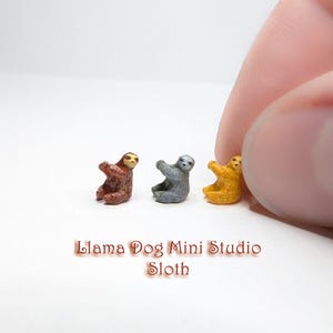 May include: Three miniature sloth figurines in brown, grey, and yellow. The sloths are posed in a sitting position with their arms raised. The figurines are arranged in a row on a white surface. The text "Llama Dog Mini Studio Sloth" is printed below the figurines.