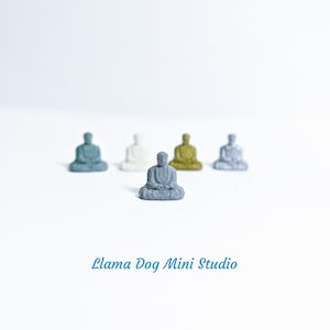 May include: Five miniature Buddha statues in various colours, including grey, white, green, gold, and silver, are arranged in a row on a white surface. The statues are all in a seated meditation pose. The text "Llama Dog Mini Studio" is printed below the statues.