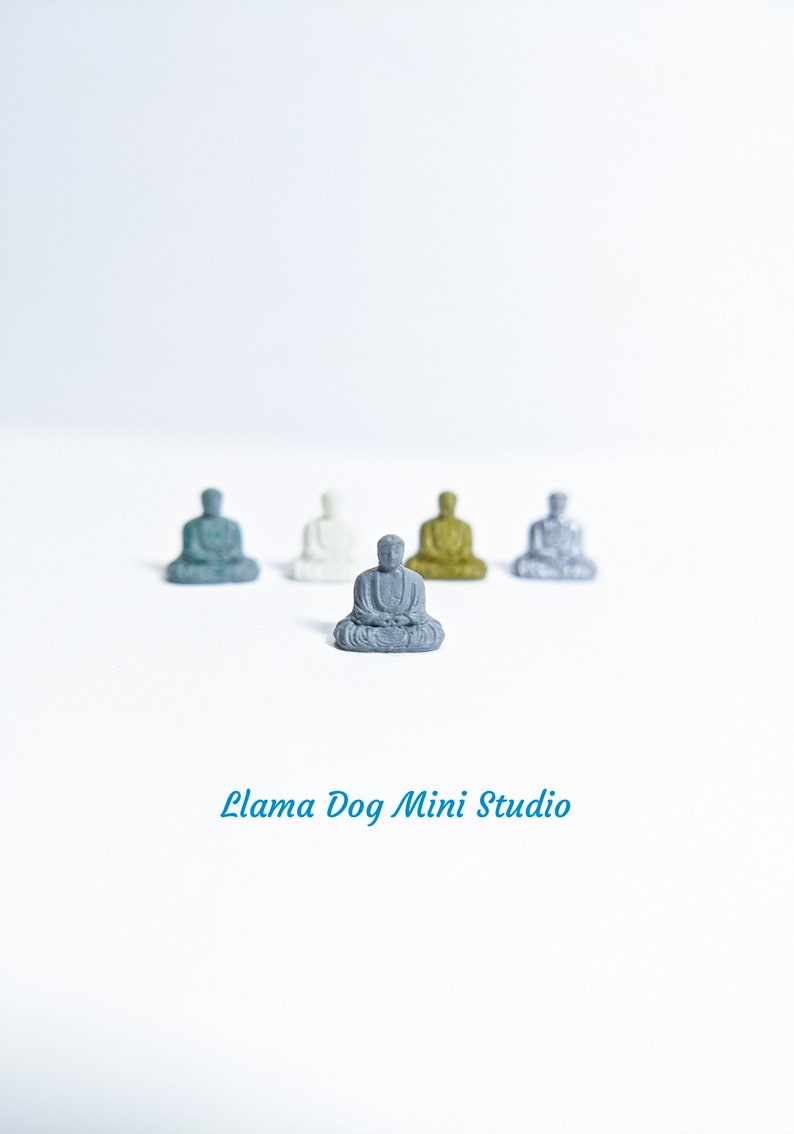 May include: Five miniature Buddha statues in various colours, including grey, white, green, gold, and silver, are arranged in a row on a white surface. The statues are all in a seated meditation pose. The text "Llama Dog Mini Studio" is printed below the statues.