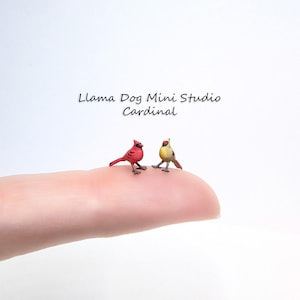 May include: Two miniature cardinal figurines, one red and one yellow, are perched on a finger. The red cardinal is facing left and the yellow cardinal is facing right. The text "Llama Dog Mini Studio Cardinal" is visible above the figurines.