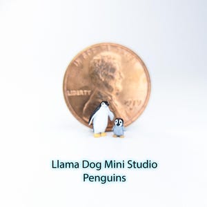 以下が含まれることがあります： 2匹のミニチュアペンギンのフィギュア。1匹は大人、もう1匹は子供で、ペニーの前に立っています。大人のペンギンは白と黒で、小さい方はグレーです。「Llama Dog Mini Studio Penguins」というテキストがペンギンの下にあります。