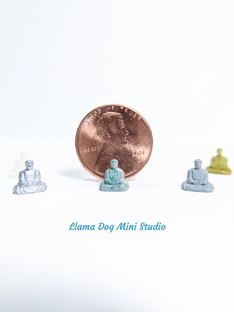 May include: Four miniature Buddha statues, one green, one silver, and two grey, are arranged on a white surface. A copper penny with the inscription "In God We Trust" and "Liberty" is in the centre of the image. The text "Llama Dog Mini Studio" is at the bottom of the image.