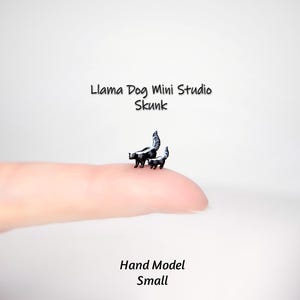May include: Two miniature skunk figurines, one adult and one juvenile, are perched on a fingertip. The skunks are black and white, with detailed markings. The text "Llama Dog Mini Studio Skunk" and "Hand Model Small" are also visible.