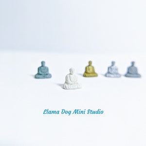 May include: A white miniature Buddha statue sits in the centre of a white surface, surrounded by four other miniature Buddha statues in various colours, including gold, silver, and blue.
