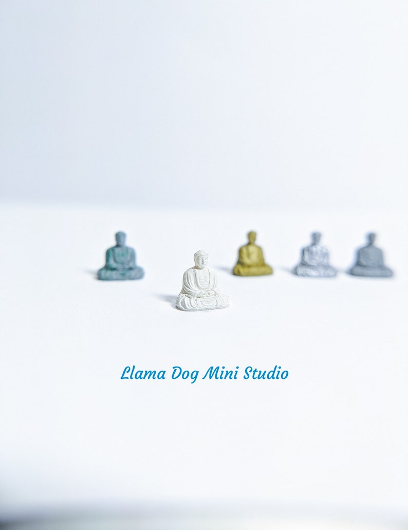 May include: A white miniature Buddha statue sits in the centre of a white surface, surrounded by four other miniature Buddha statues in various colours, including gold, silver, and blue.