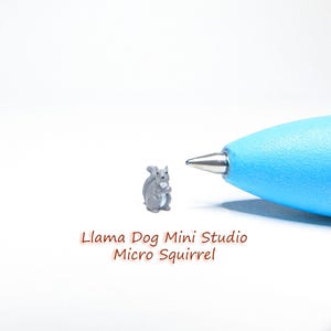 Peut inclure: Une petite figurine de  écureuil gris, de la taille d'un stylo, est présentée sur une surface blanche. La figurine tient une noix dans ses pattes. Le texte "Llama Dog Mini Studio Micro Squirrel" est écrit sous la figurine.