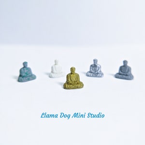 May include: Five miniature Buddha statues in various colours, including gold, silver, and grey, sitting on a white surface. The gold Buddha is in the centre of the image.