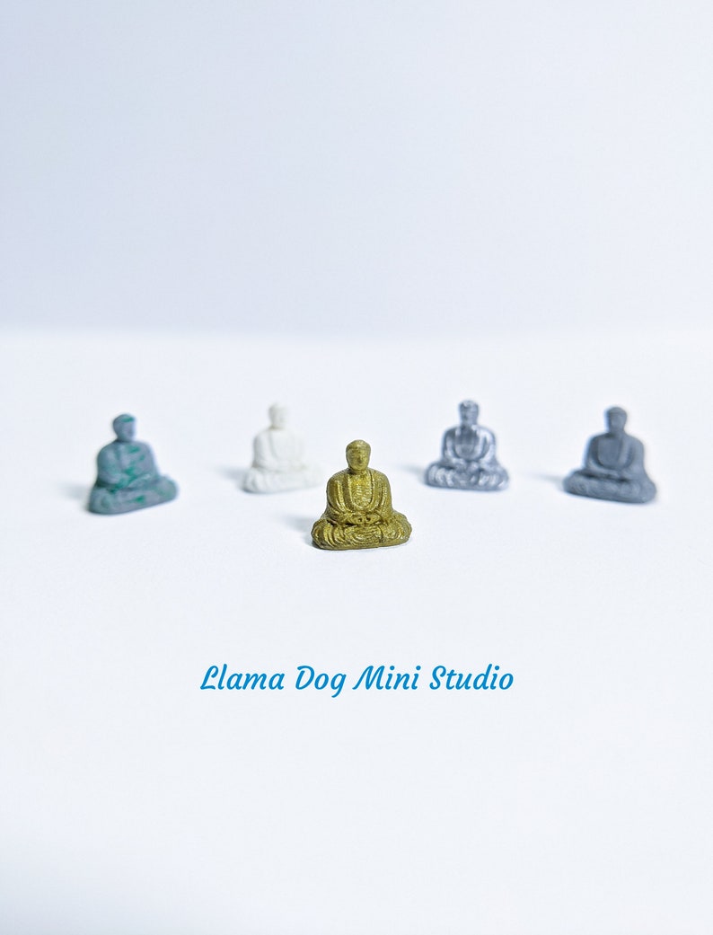 May include: Five miniature Buddha statues in various colours, including gold, silver, and grey, sitting on a white surface. The gold Buddha is in the centre of the image.