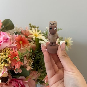May include: A small, brown figurine of a cartoon character with large eyes and a textured body. The figurine is held in front of a colorful bouquet of pink, orange, and yellow flowers. The figurine is approximately 2 inches tall.