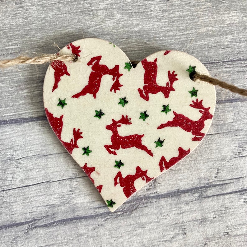 May include: A heart-shaped wooden ornament featuring a cream-colored base with a repeating pattern of red reindeer and small green stars. The ornament has two holes at the top, threaded with a thin brown twine string for hanging purposes.