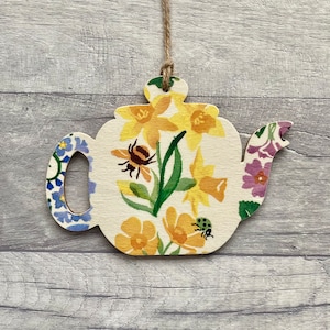May include: A wooden teapot ornament with a floral design featuring yellow daffodils, green leaves, a bee, and a ladybug. The teapot is painted on a white background and has a blue and white patterned handle.