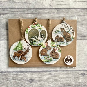 May include: Four circular wooden ornaments featuring illustrations of a deer family, badgers, foxes, and rabbits in snowy landscapes. Each ornament has a jute twine loop for hanging. A small round sticker with a dog illustration and the text Krafty Bob.
