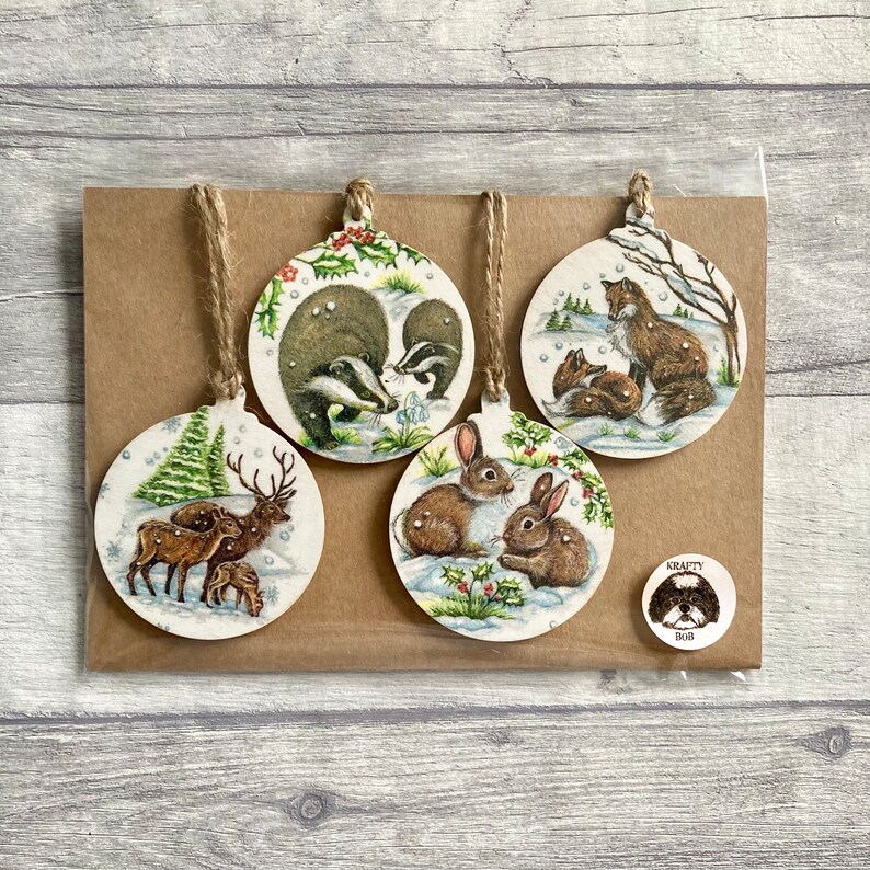 May include: Four circular wooden ornaments featuring illustrations of a deer family, badgers, foxes, and rabbits in snowy landscapes. Each ornament has a jute twine loop for hanging. A small round sticker with a dog illustration and the text Krafty Bob.