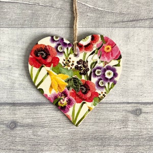 A wooden heart-shaped ornament with a floral pattern. The ornament is decorated with a variety of flowers in shades of red, pink, purple, yellow, and green. The ornament is hanging with natural twine.