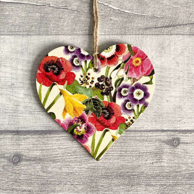 A wooden heart-shaped ornament with a floral pattern. The ornament is decorated with a variety of flowers in shades of red, pink, purple, yellow, and green. The ornament is hanging with natural twine.
