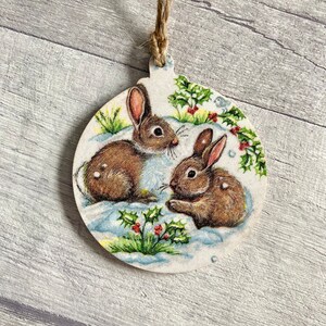May include: A round wooden ornament featuring a printed illustration of two brown rabbits sitting in white snow. The design includes green holly leaves with small red berries and sprigs of grass. A piece of brown twine is attached to the top loop.