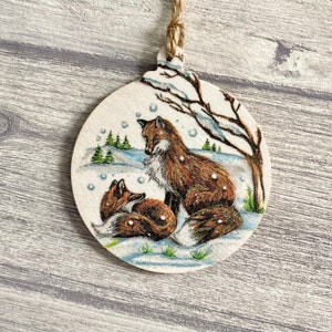 May include: A round wooden ornament featuring a printed illustration of two foxes in a snowy landscape. One fox sits upright while the other rests nearby. The scene includes a bare tree, small evergreen trees, and falling snowflakes. A jute twine loop is attached.